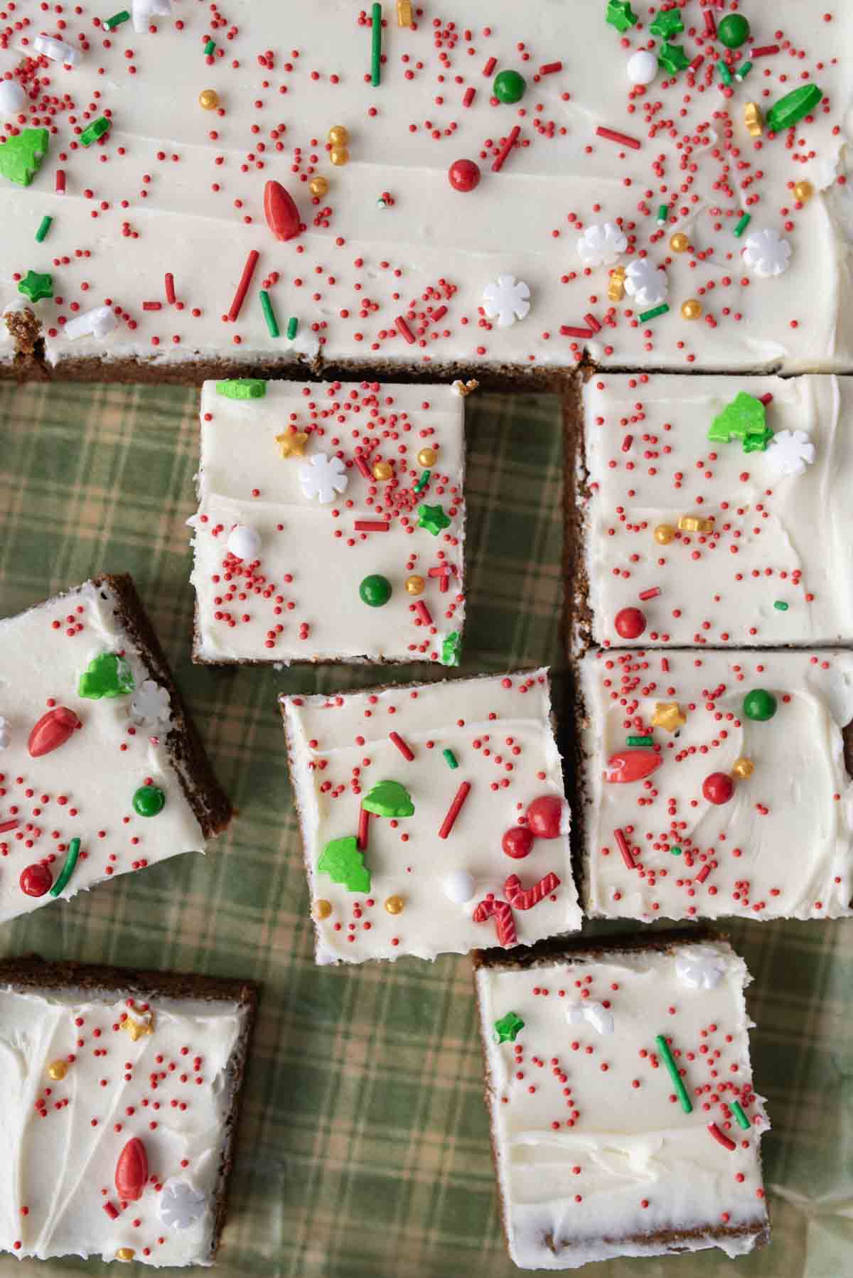Frosted gingerbread bars cut into squares, topped with white icing and festive red, green, and gold holiday sprinkles, are arranged on a green plaid surface.