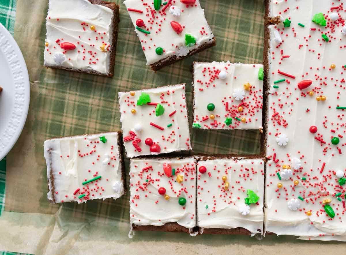 Frosted holiday cookie bars cut into squares, topped with white icing and festive red, green, and white sprinkles, on parchment paper over a plaid surface.
