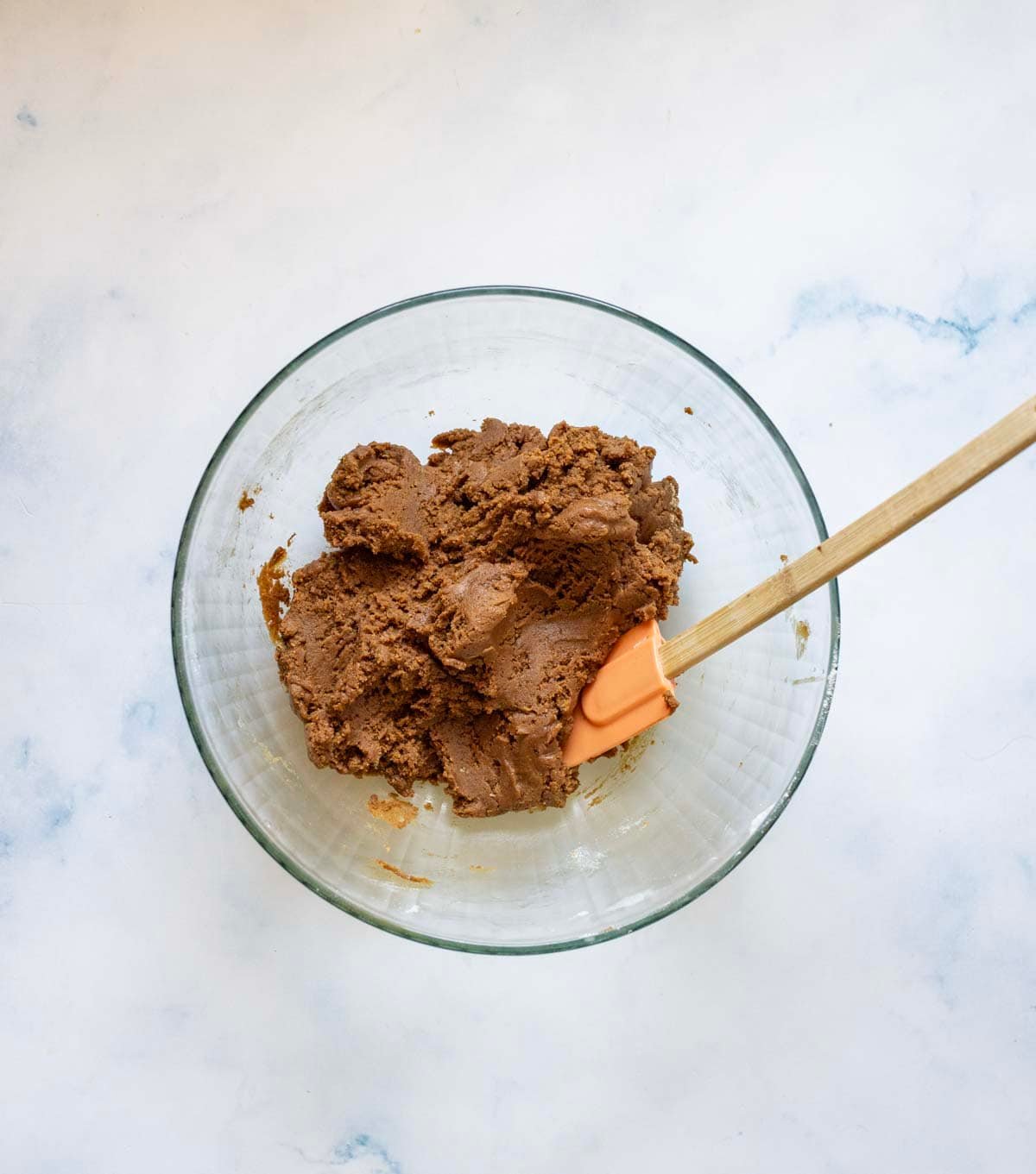 A glass bowl filled with chocolate cookie dough sits on a white surface. An orange spatula with a wooden handle rests in the dough.