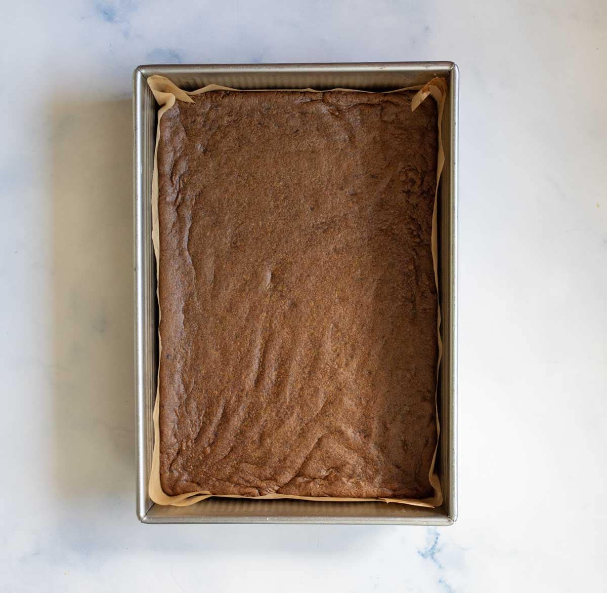 A rectangular pan lined with parchment paper, filled with a freshly baked, uncut chocolate brownie, sits on a light-colored surface.