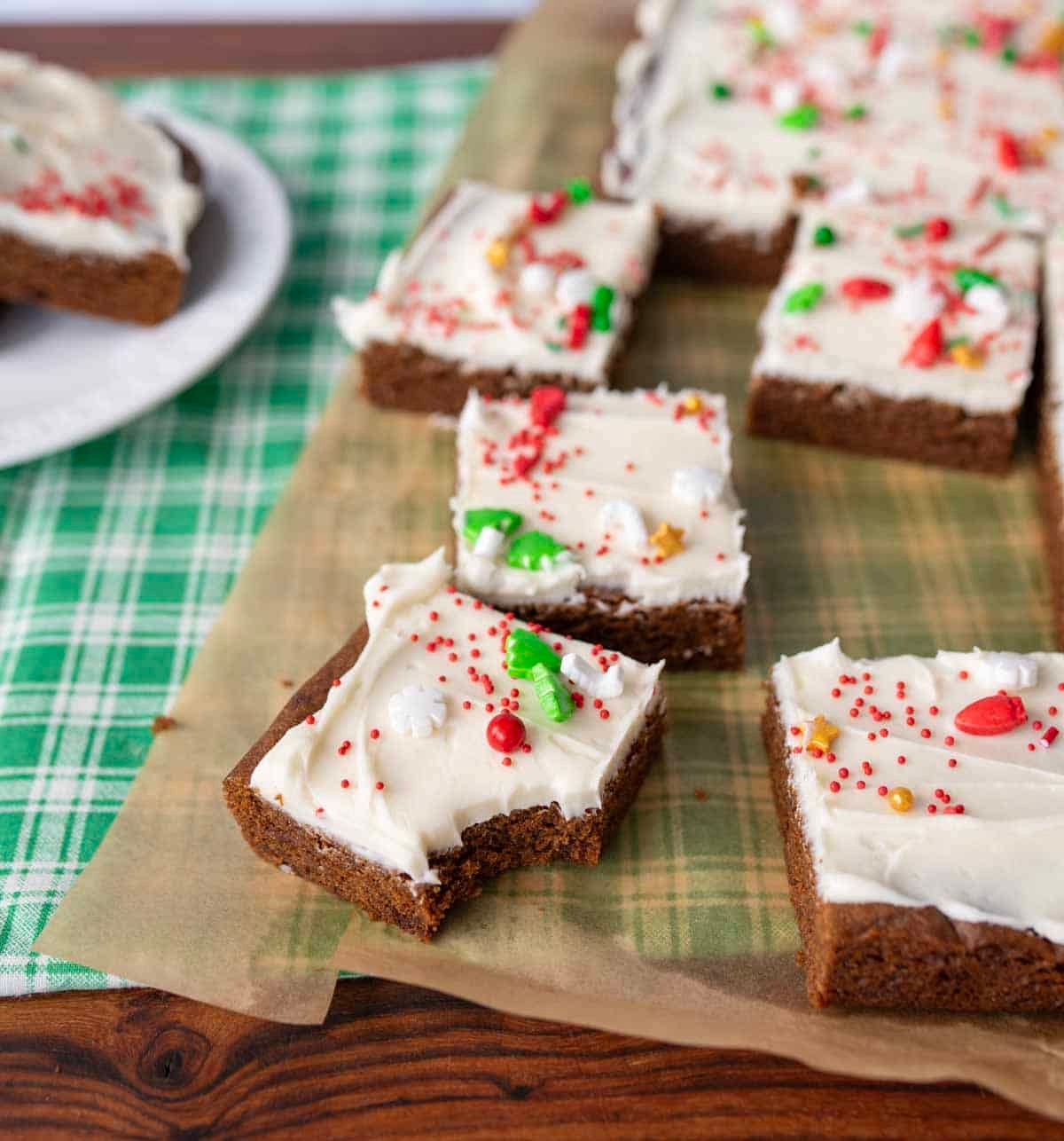 Frosted gingerbread bars with colorful holiday sprinkles are arranged on parchment paper. One bar in the foreground has a bite taken out of it. A green plaid cloth is partially visible underneath.