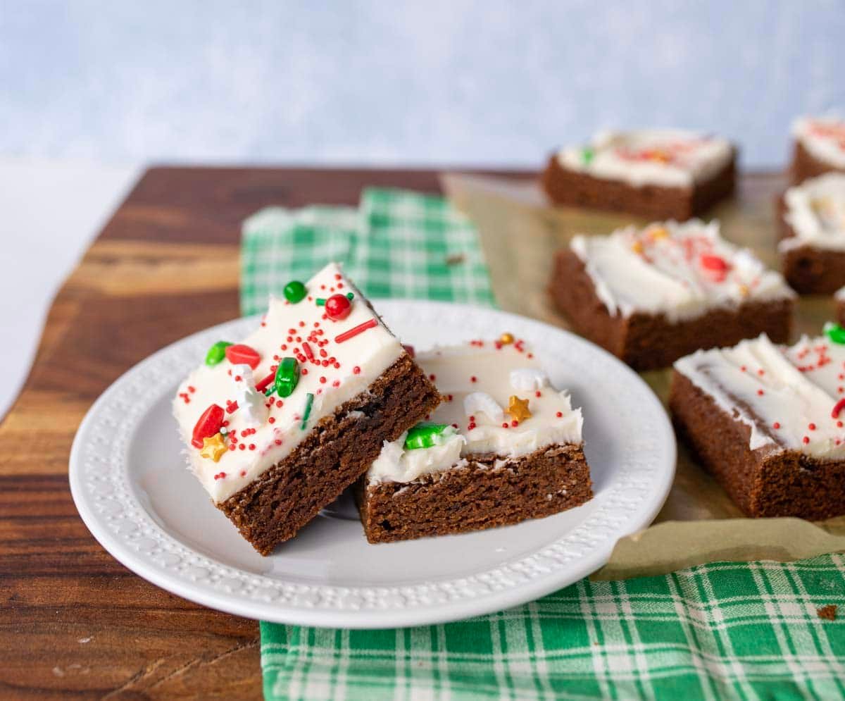 Two chocolate brownies with white frosting and colorful sprinkles are stacked on a white plate, with more frosted brownies in the background on a wooden surface and green plaid cloth.