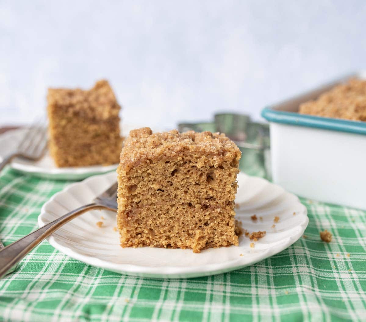A piece of coffee cake with crumb topping sits on a white plate with a fork, placed on a green plaid cloth. More cake and a baking dish are blurred in the background.