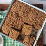 A pan of cinnamon gingerbread coffee cake, sliced into squares, with several pieces slightly lifted. The cake has a crumbly streusel topping and sits on a green checkered cloth.