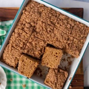 A pan of cinnamon gingerbread coffee cake, sliced into squares, with several pieces slightly lifted. The cake has a crumbly streusel topping and sits on a green checkered cloth.