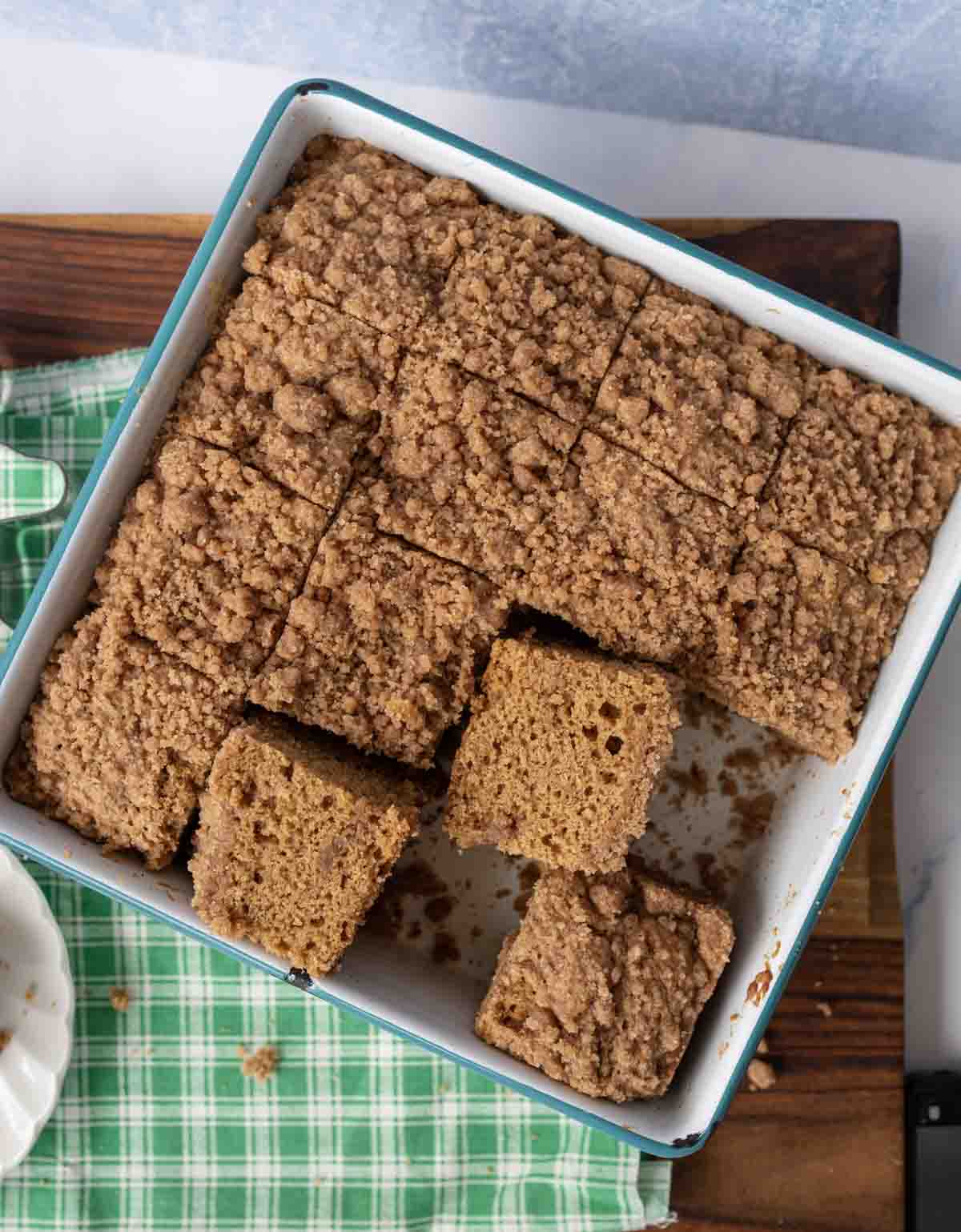A pan of cinnamon gingerbread coffee cake, sliced into squares, with several pieces slightly lifted. The cake has a crumbly streusel topping and sits on a green checkered cloth.