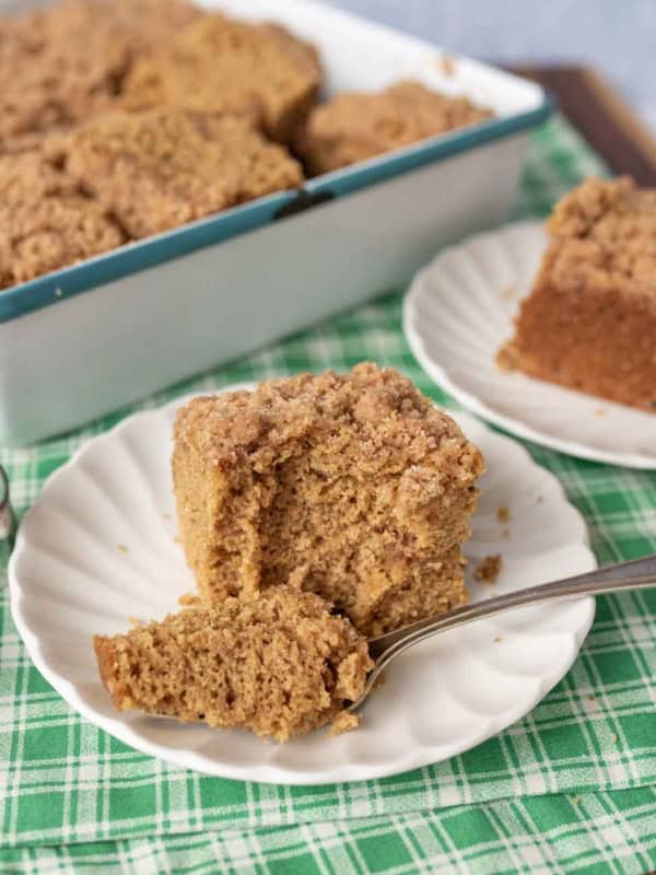 A slice of crumb-topped gingerbread coffee cake on a white plate with a fork, with a baking dish of the same cake and another plated slice in the background, all on a green checked cloth.