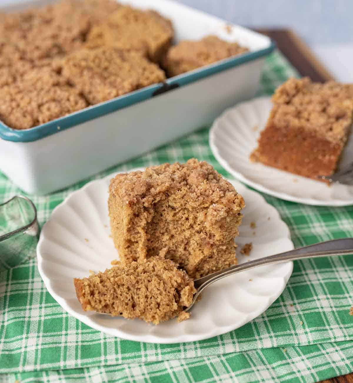 A slice of crumb-topped gingerbread coffee cake on a white plate with a fork, with a baking dish of the same cake and another plated slice in the background, all on a green checked cloth.