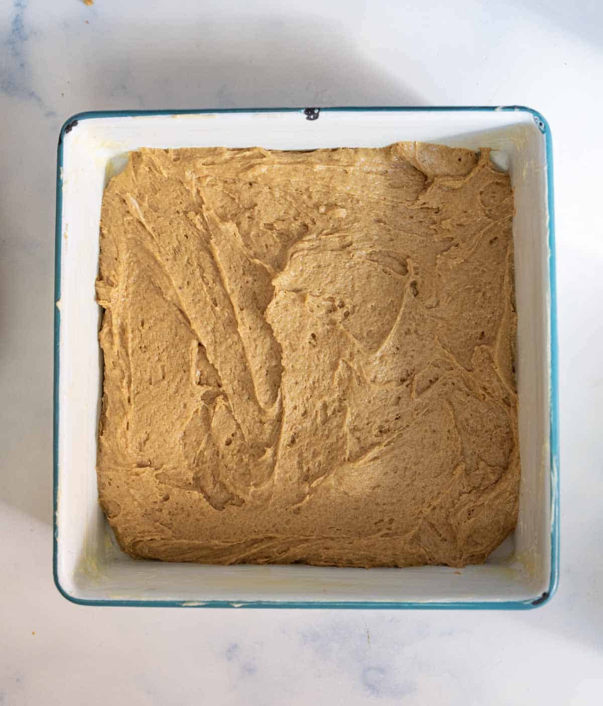 A square baking dish filled with an even layer of light brown batter, ready to be baked, sits on a white countertop.