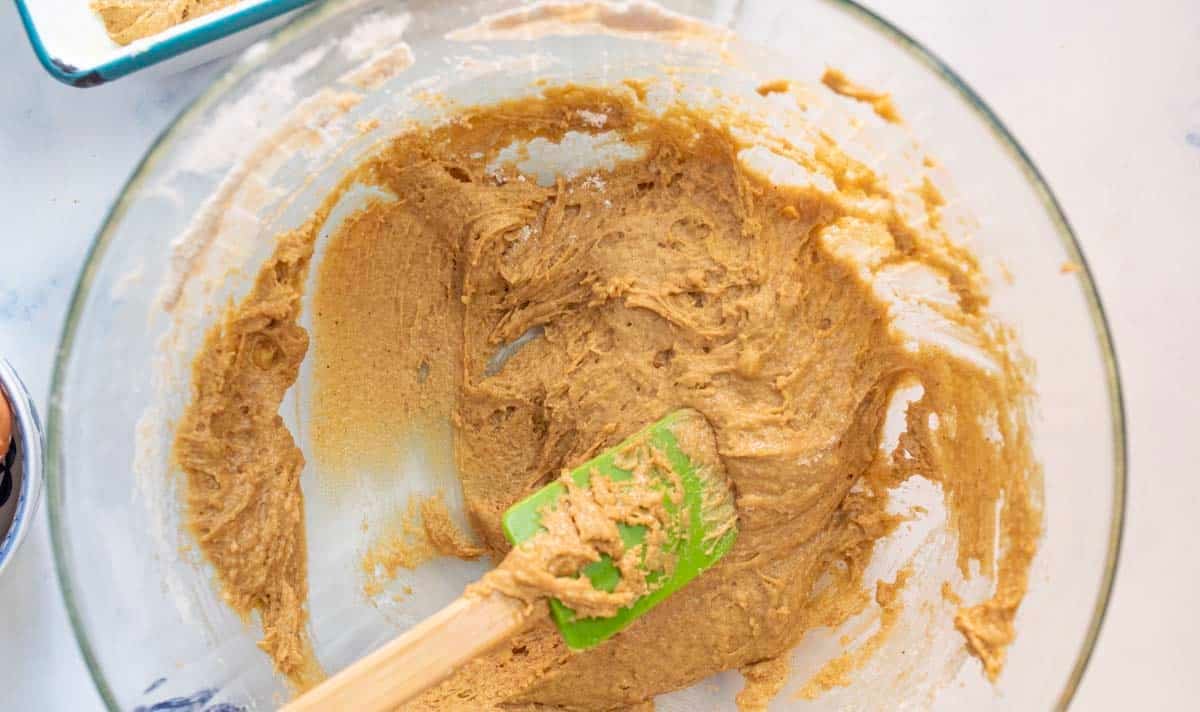 A glass mixing bowl containing cookie dough being stirred with a green spatula. The dough is light brown and partially mixed, with some residue on the sides of the bowl.