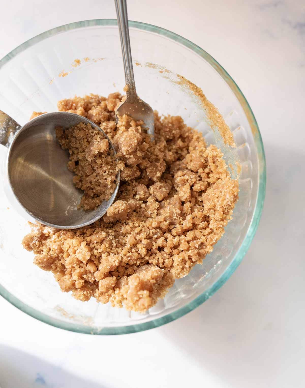 A glass bowl containing a crumbly graham cracker mixture with a metal measuring cup and a fork resting inside. The bowl sits on a light-colored surface.