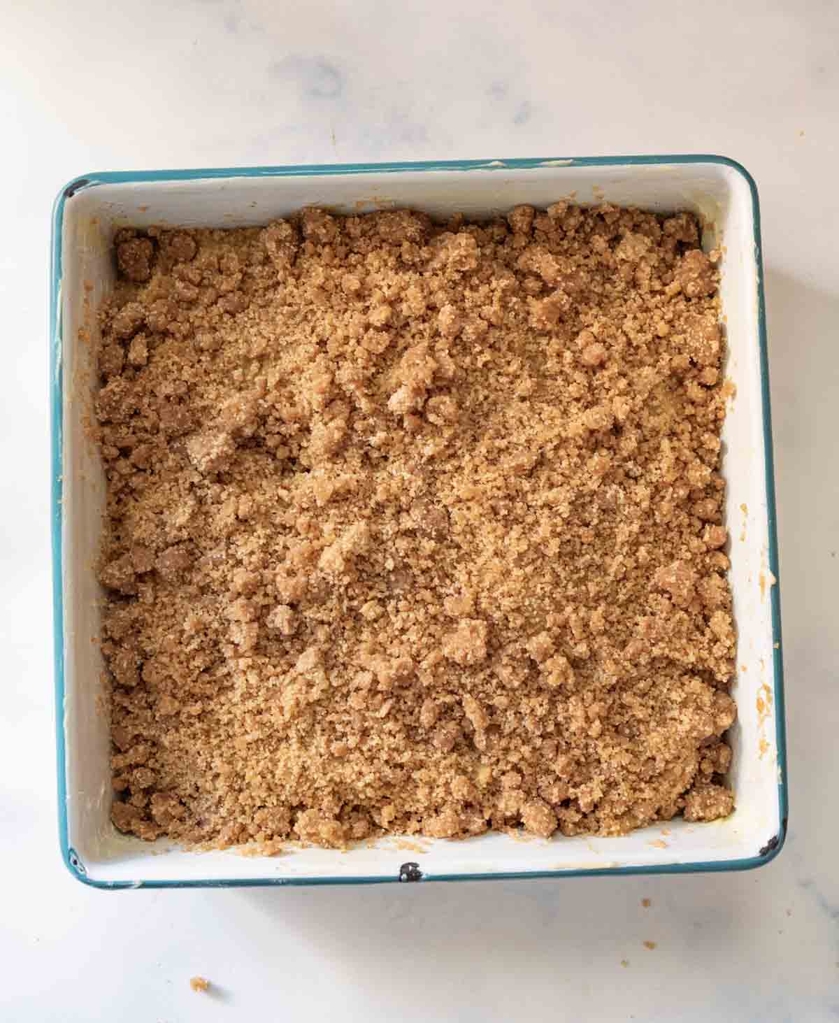 A square baking dish filled with a golden brown crumb topping, likely for a coffee cake or crumble dessert, set on a light marble surface.