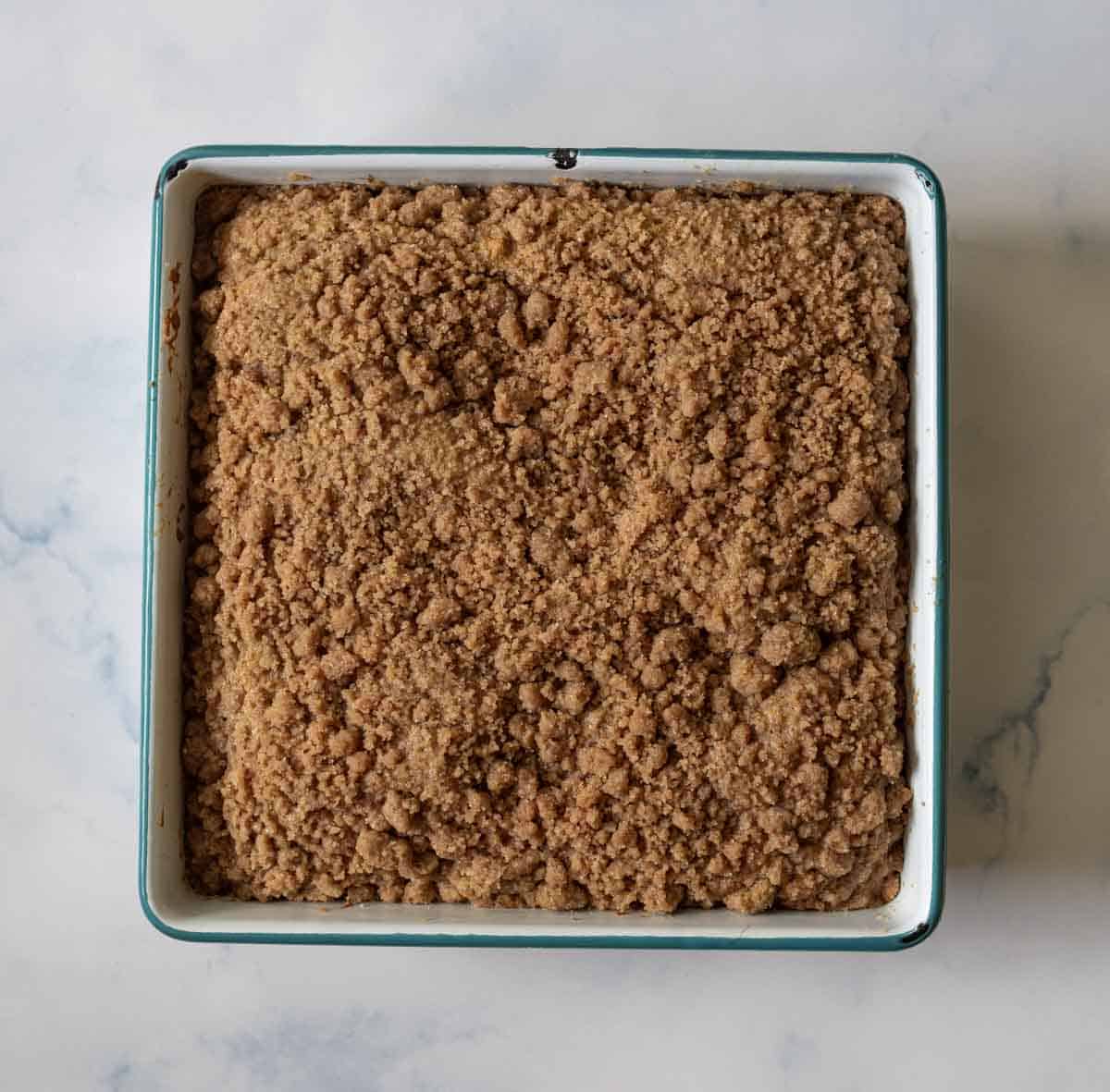 A square baking dish filled with coffee cake topped with a thick, crumbly cinnamon streusel, sitting on a light marble surface.