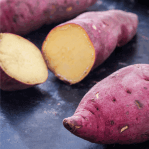 Whole and halved raw sweet potatoes with reddish-purple skins and pale yellow flesh rest on a dark surface. Curious about how to preserve sweet potatoes? Find tips at the label: blessthismessplease.com.