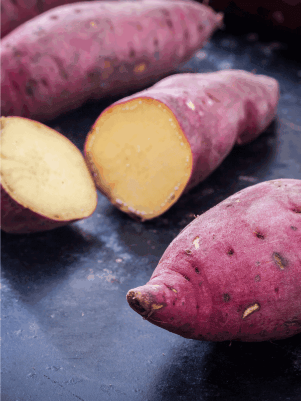 Whole and halved raw sweet potatoes with reddish-purple skins and pale yellow flesh rest on a dark surface. Curious about how to preserve sweet potatoes? Find tips at the label: blessthismessplease.com.