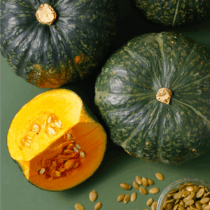 Three whole green kabocha squash, one cut open to reveal yellow flesh and seeds, with a small bowl of pumpkin seeds nearby on a green surface. Learn how to preserve winter squash at blessthismessplease.com.