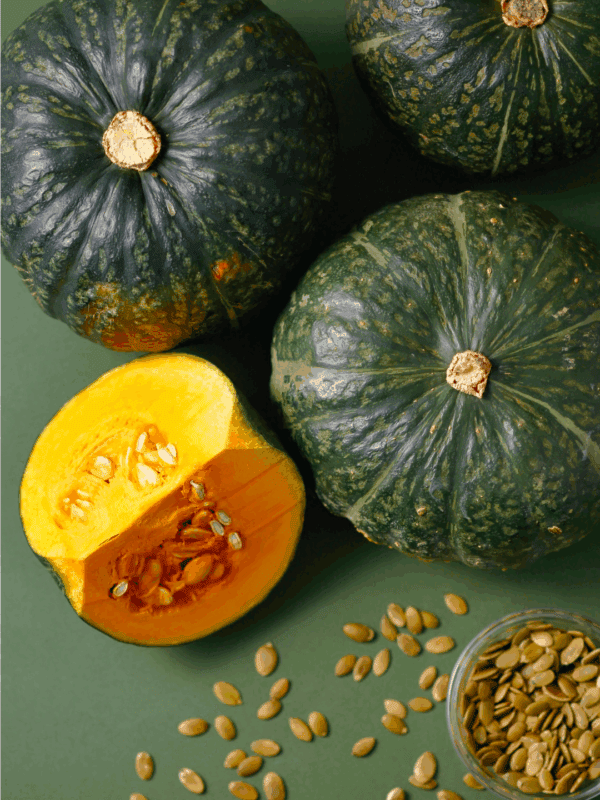 Three whole green kabocha squash, one cut open to reveal yellow flesh and seeds, with a small bowl of pumpkin seeds nearby on a green surface. Learn how to preserve winter squash at blessthismessplease.com.