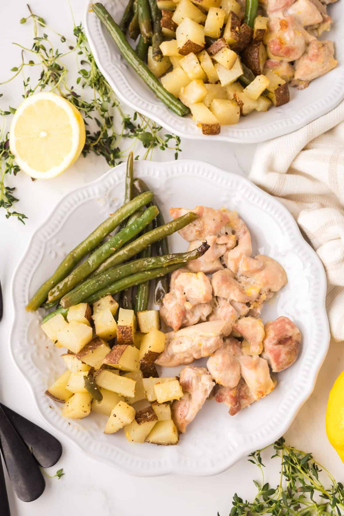 Two white plates with roasted green beans, diced potatoes, and pieces of cooked chicken. Fresh thyme and lemon halves are on the table as garnishes. A beige cloth napkin is nearby.