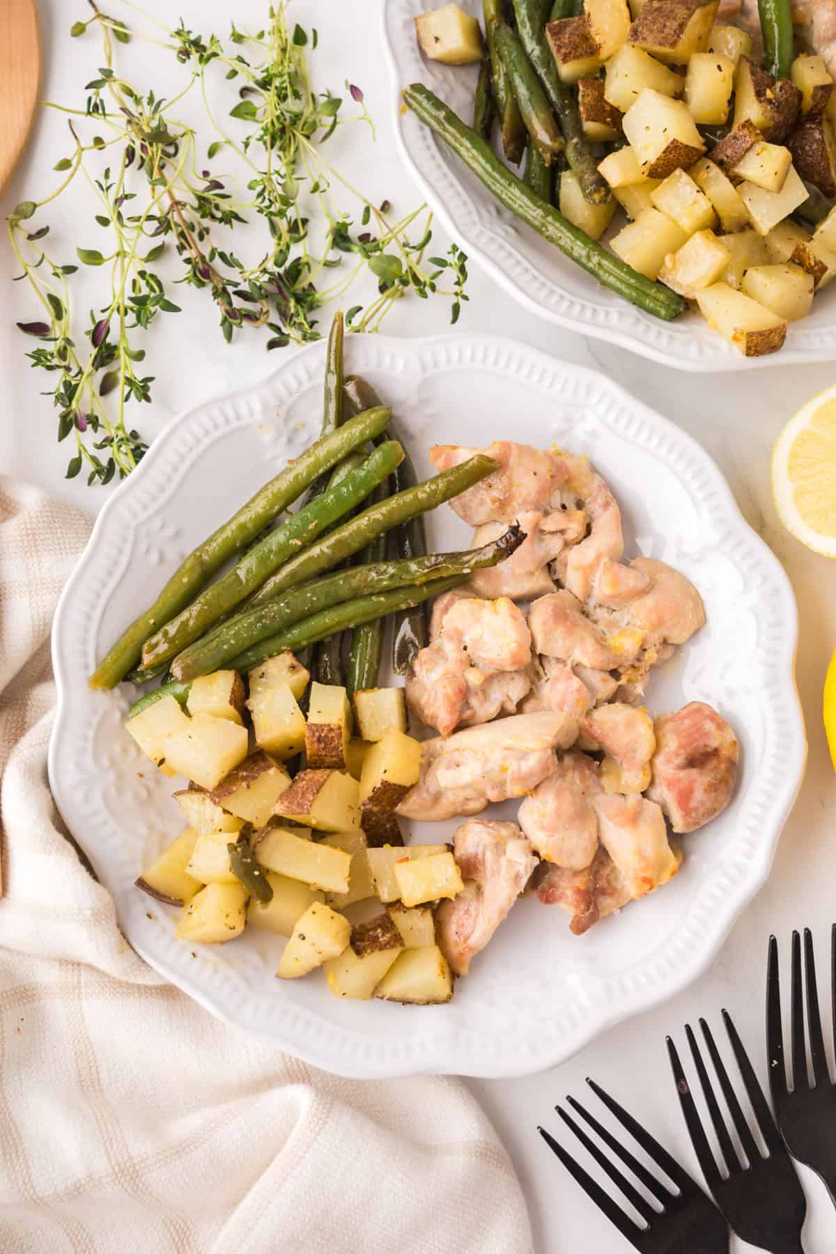 A white plate with roasted chicken pieces, green beans, and diced potatoes. Fresh thyme, lemon slices, and another plate of food are nearby on a white surface. Black utensils and a cloth napkin are also visible.