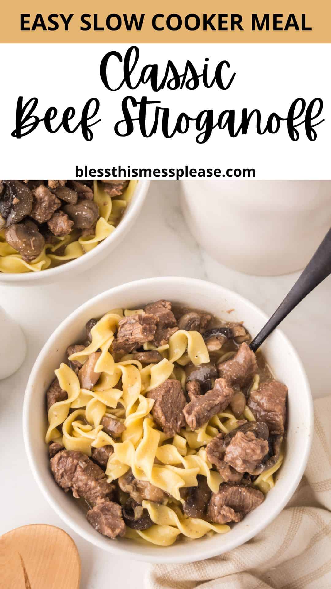 A bowl of slow cooker beef stroganoff with tender beef chunks and egg noodles sits on a white surface. A spoon rests in the bowl, while another smaller bowl with stroganoff is partially visible in the background.