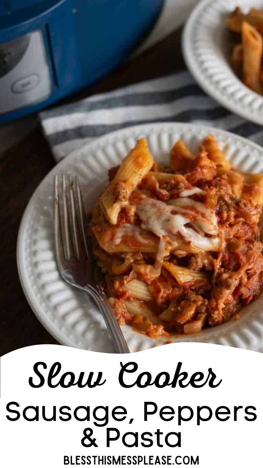 A white plate with baked pasta topped with melted cheese, slow cooker sausage, and peppers, with a fork on the side. A slow cooker and another plate are partially visible in the background.