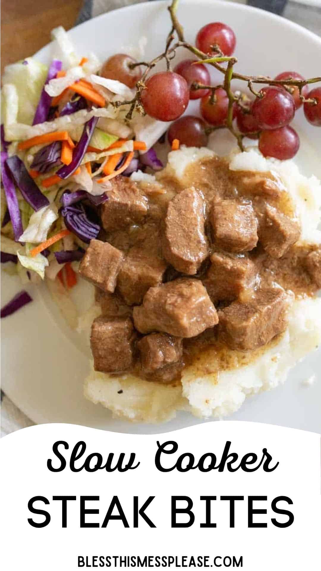 A plate with slow cooker steak bites over mashed potatoes, served alongside a fresh salad and red grapes. Text reads: Slow Cooker Steak Bites. blessthismessplease.com.