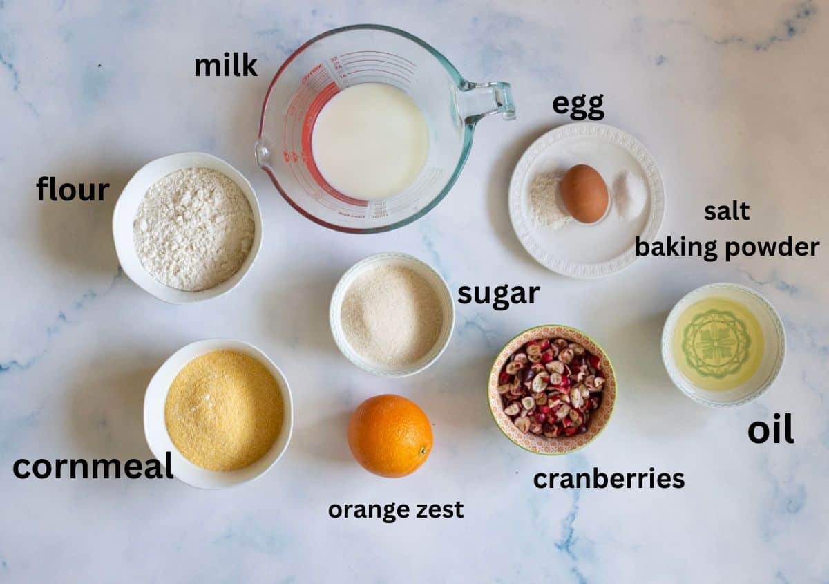Baking ingredients arranged on a table: flour, cornmeal, milk in a measuring cup, an egg, salt, baking powder, sugar, oil, cranberries, and an orange labeled as orange zest.