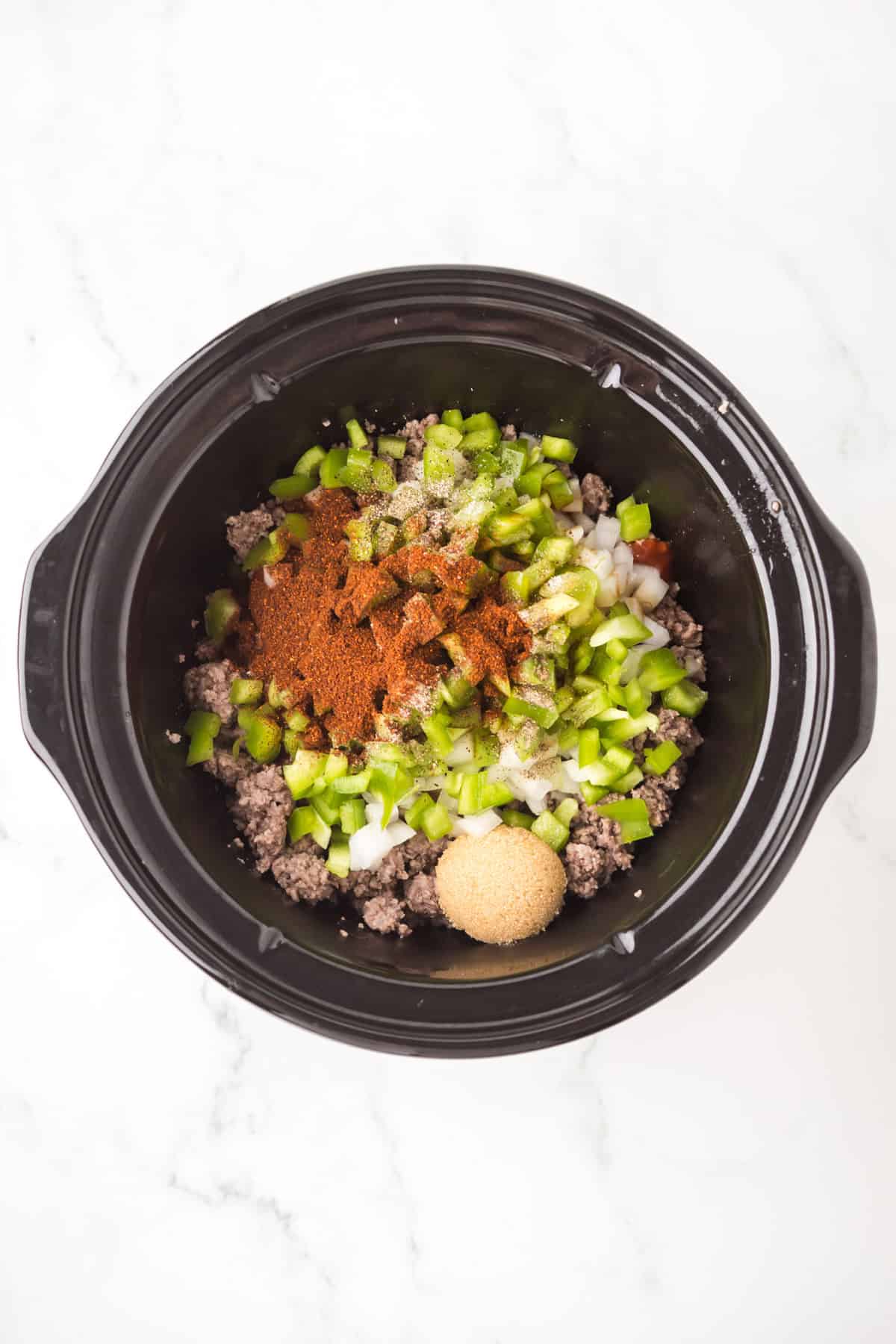 A crock pot sloppy joes mixture with ground beef, chopped green bell peppers, diced onions, brown sugar, and spices sits on a white marble surface.