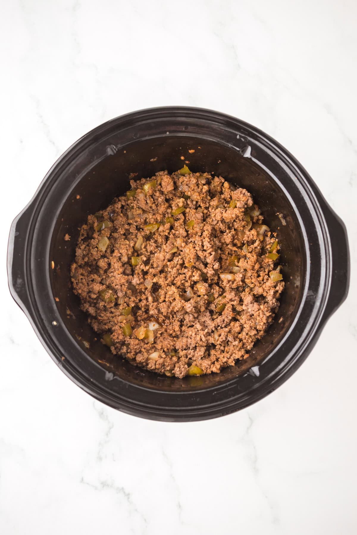 Ground beef mixed with chopped onions and green peppers in a black slow cooker, perfect for making crock pot sloppy joes, viewed from above on a white marble background.