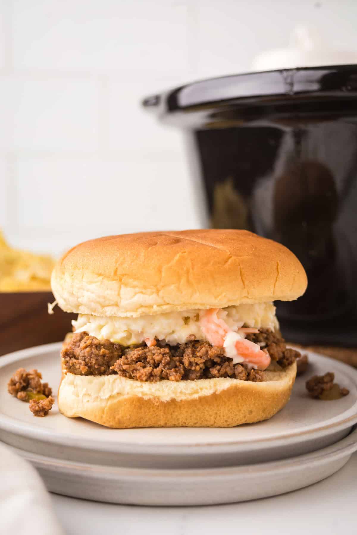 A crock pot sloppy joes sandwich topped with coleslaw sits on a plate, with a slow cooker and tortilla chips in the background.
