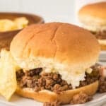 Crock pot sloppy joes sandwich with ground beef filling and a dollop of coleslaw is served on a plate with potato chips. Another crock pot sloppy joes sandwich and more chips are visible in the background.