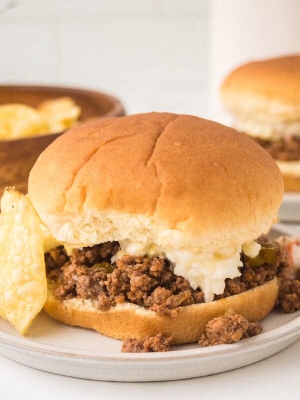 Crock pot sloppy joes sandwich with ground beef filling and a dollop of coleslaw is served on a plate with potato chips. Another crock pot sloppy joes sandwich and more chips are visible in the background.