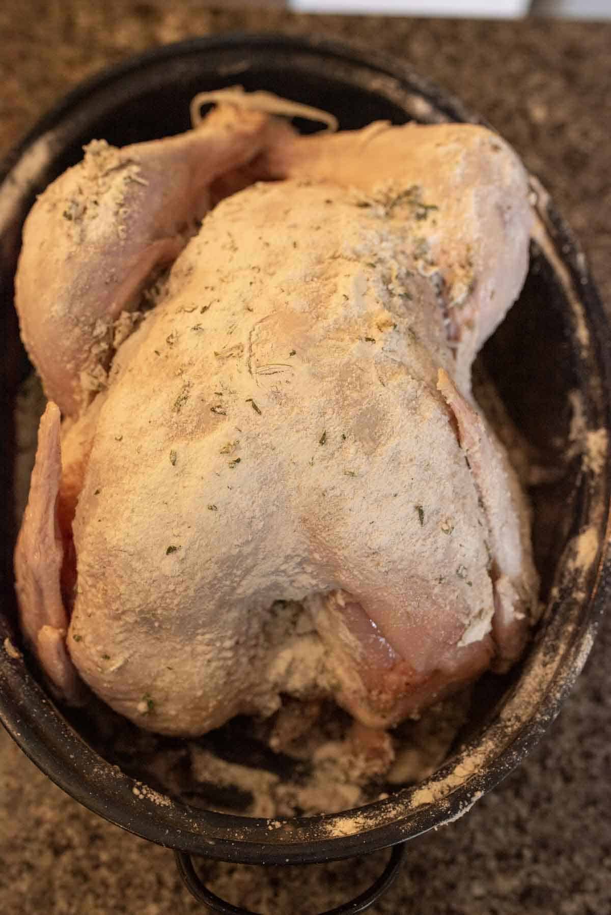 A whole raw chicken in a roasting pan, coated with a layer of flour and herbs, sitting on a kitchen countertop.