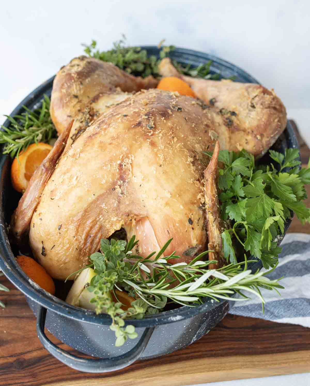 Roast turkey in a large pan, garnished with fresh herbs like rosemary and parsley, and halved oranges; set on a wooden surface with a striped towel nearby.