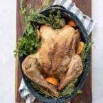 A dry brine turkey, roasted whole and garnished with fresh herbs and orange slices, sits in a large roasting pan on a wooden board with a striped kitchen towel underneath.