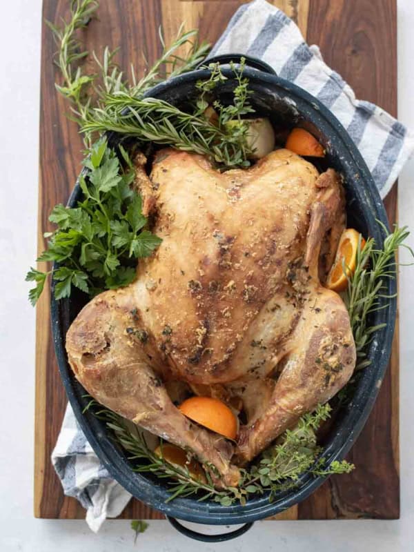 A dry brine turkey, roasted whole and garnished with fresh herbs and orange slices, sits in a large roasting pan on a wooden board with a striped kitchen towel underneath.