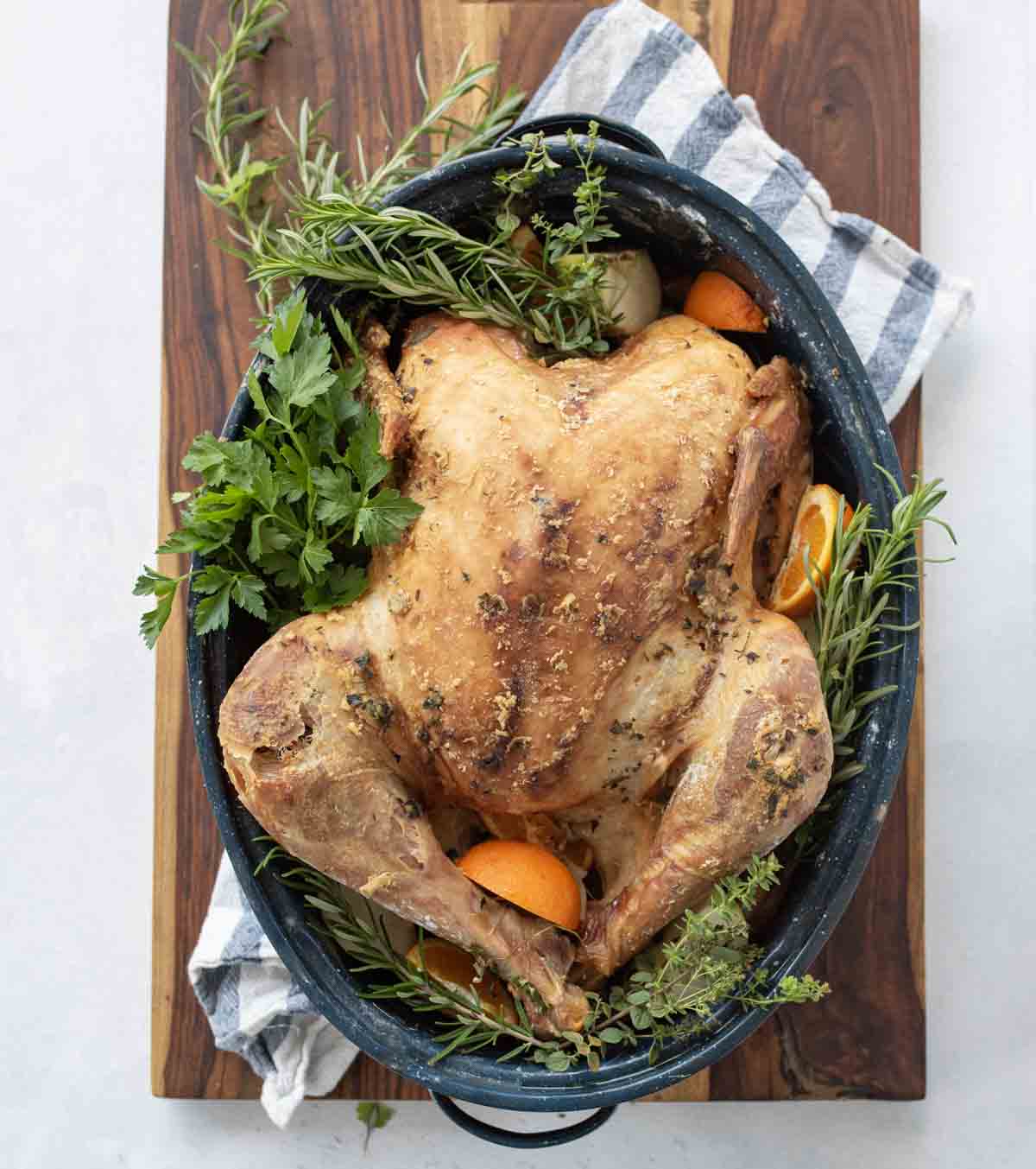 A dry brine turkey, roasted whole and garnished with fresh herbs and orange slices, sits in a large roasting pan on a wooden board with a striped kitchen towel underneath.