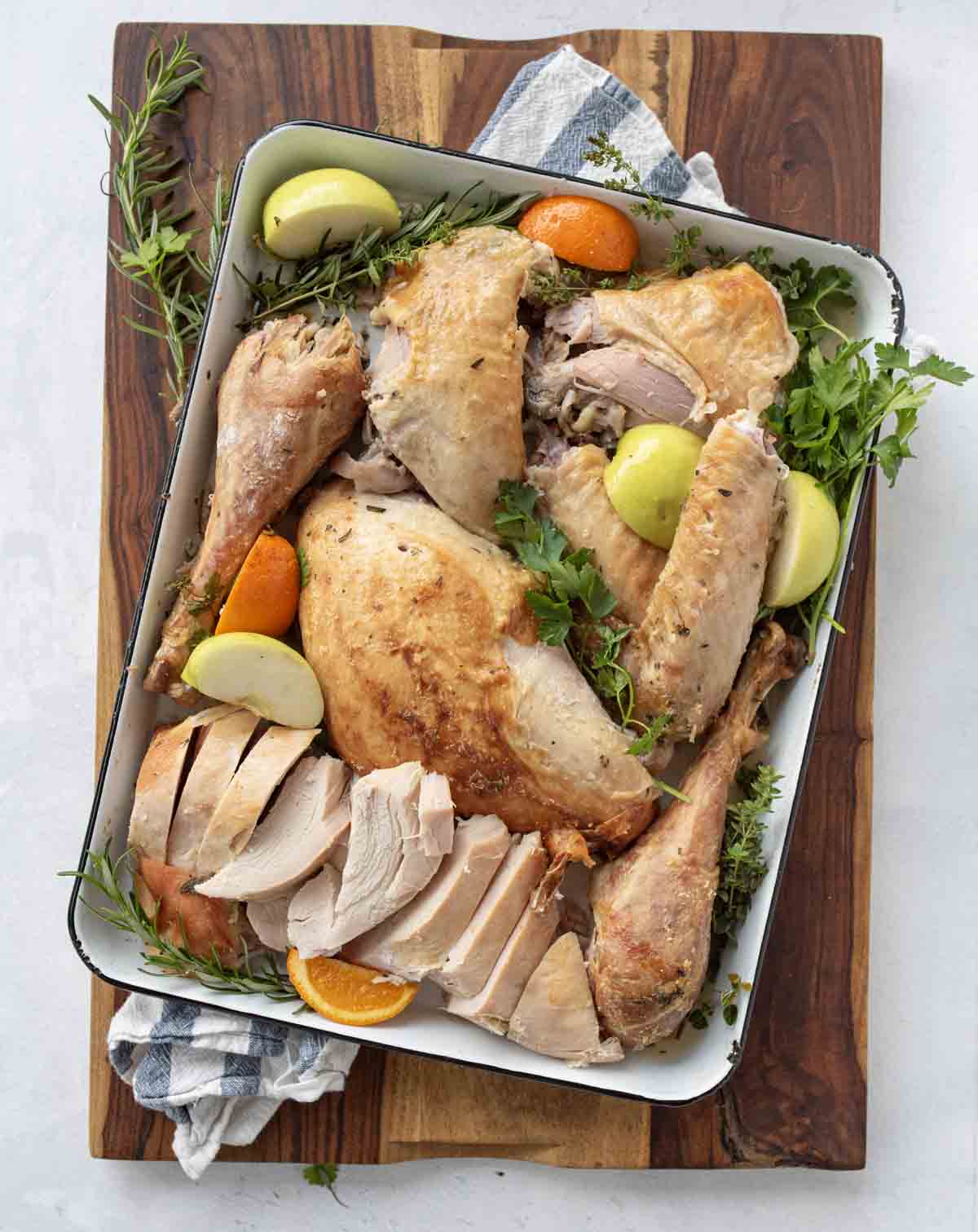 A platter of carved roasted turkey is garnished with fresh herbs, green apple slices, and orange wedges, set on a wooden cutting board with a striped kitchen towel underneath.