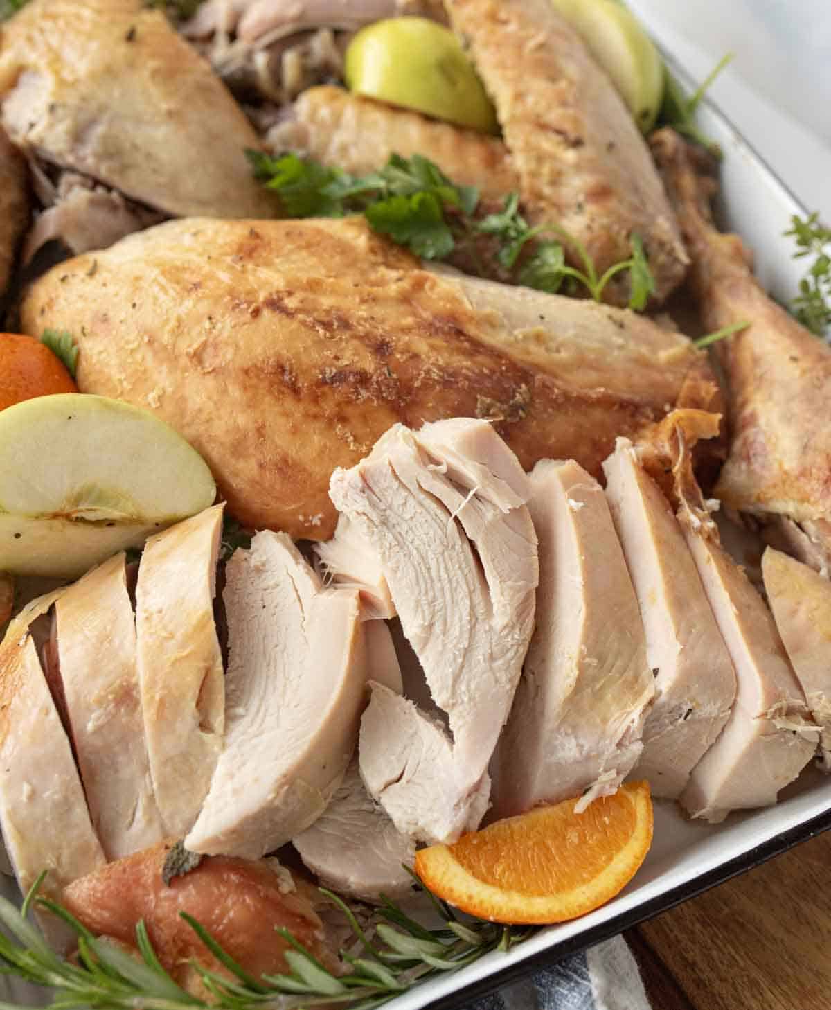 A platter of sliced roasted turkey breast, garnished with fresh herbs, apple wedges, and orange slices. The turkey is golden brown and juicy, arranged on a white serving dish.