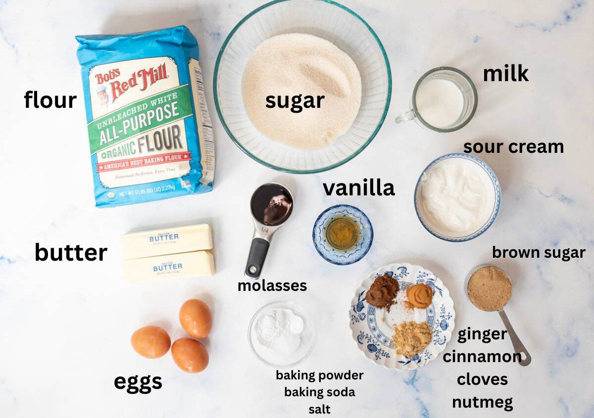 A flat lay of baking ingredients labeled: flour, sugar, milk, sour cream, vanilla, brown sugar, molasses, butter, eggs, and small bowls of baking powder, baking soda, salt, ginger, cinnamon, cloves, and nutmeg.