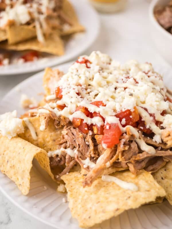 A plate of pulled pork nachos topped with diced tomatoes, melted cheese, and drizzled with a white sauce, served on a white plate. Another plate of nachos is visible in the background.