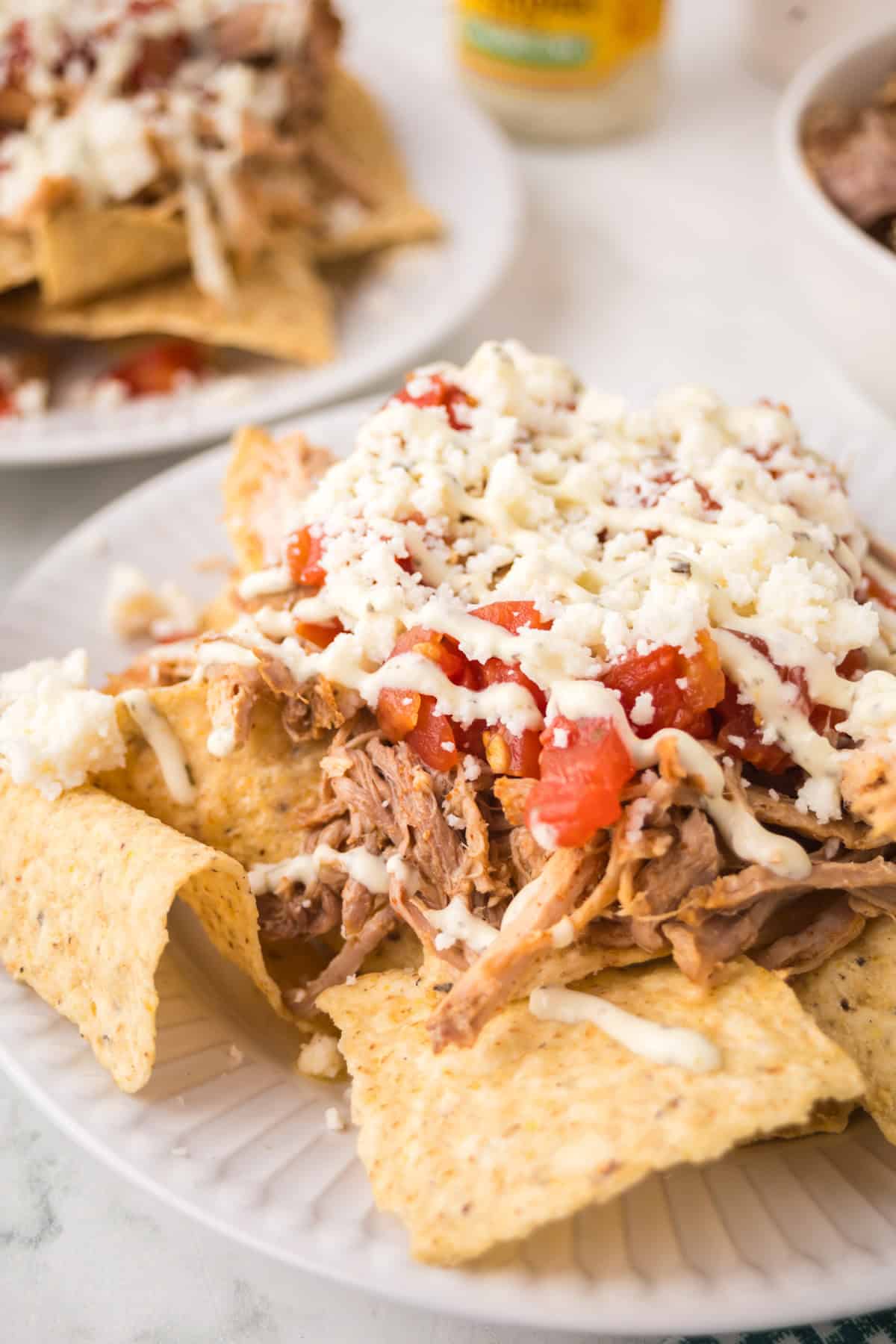 A plate of pulled pork nachos topped with diced tomatoes, melted cheese, and drizzled with a white sauce, served on a white plate. Another plate of nachos is visible in the background.