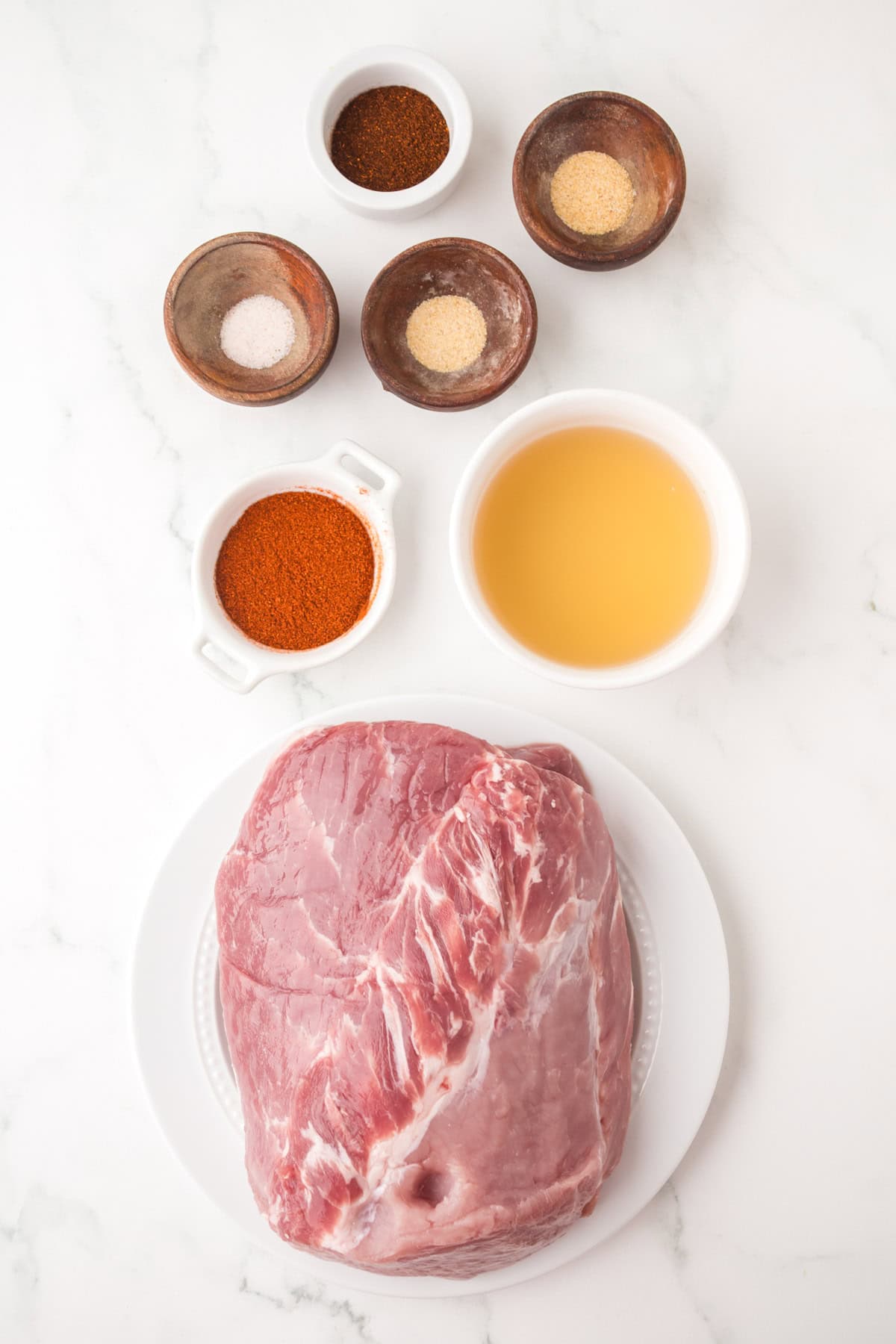 Raw pork shoulder on a white plate surrounded by small bowls of apple cider vinegar, paprika, salt, pepper, chili powder, and garlic powder—perfect for preparing flavorful pulled pork nachos on a white marble surface.