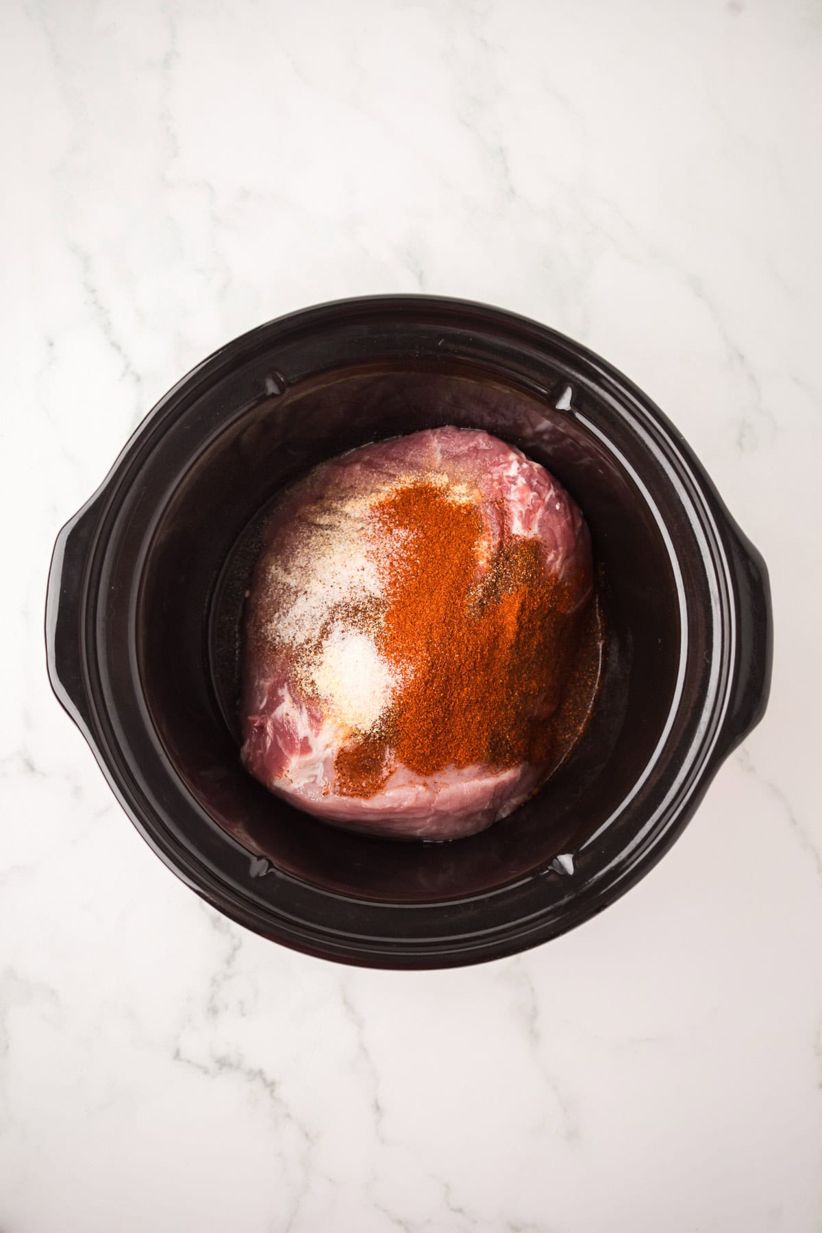 Raw pork roast topped with spices like paprika and garlic powder, ready in a black slow cooker on a white marble surface—perfect for making flavorful pulled pork nachos.