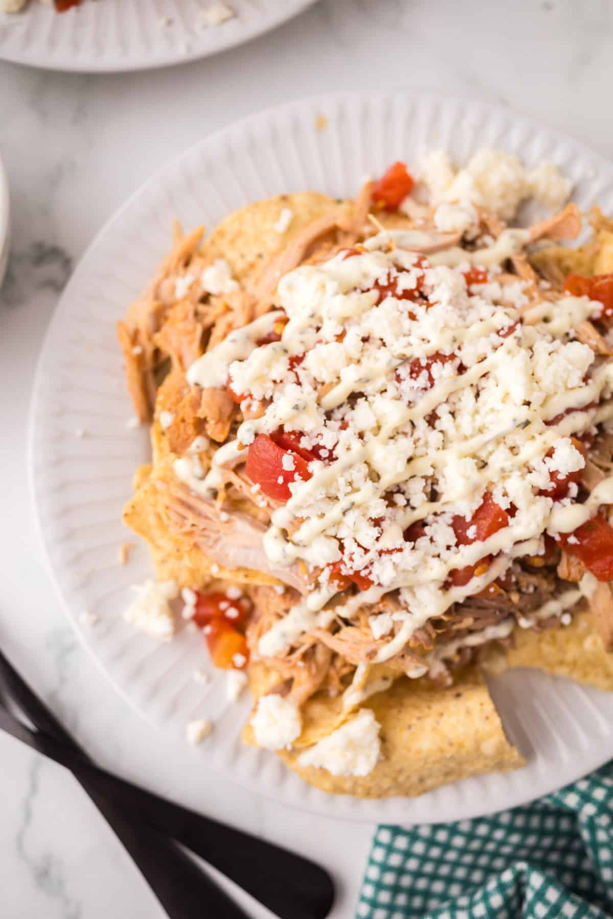 A plate of pulled pork nachos topped with diced tomatoes, crumbled cheese, and drizzled with a creamy white sauce sits on a white plate next to a fork and a green checkered napkin.