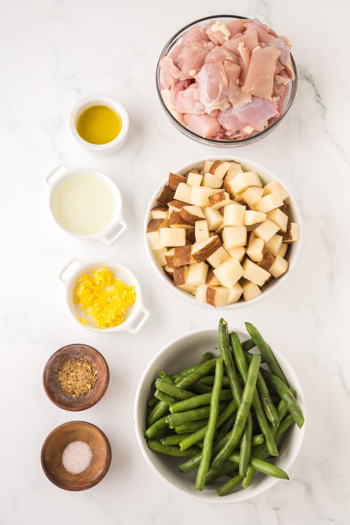 Overhead view of ingredients on a white surface: a bowl of raw chicken pieces, a bowl of chopped potatoes, a bowl of fresh green beans, minced garlic, olive oil, milk, dried herbs, and salt in small containers.
