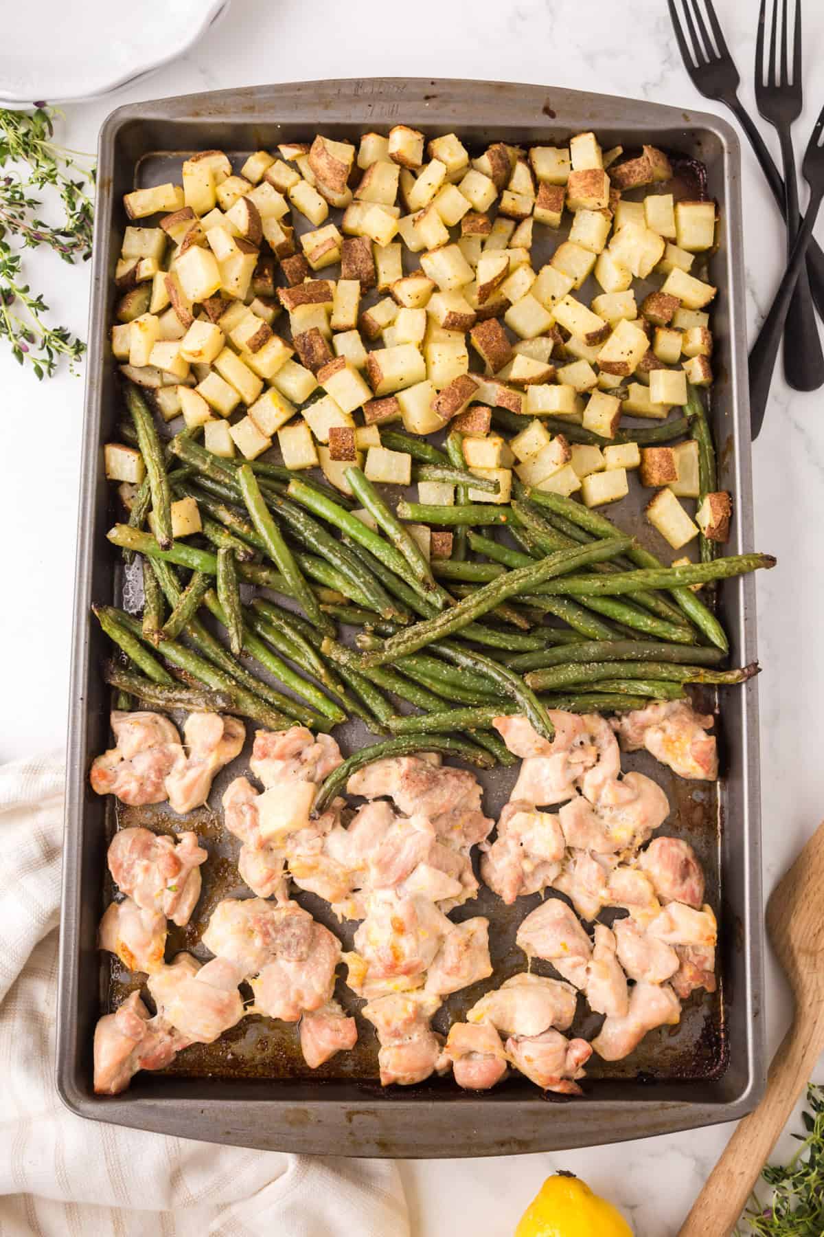 A sheet pan with three sections: roasted diced potatoes in the top left, green beans in the center, and pieces of cooked chicken in the bottom right, on a marble surface with utensils and a napkin nearby.