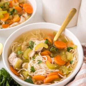 A bowl of slow cooker chicken soup with sliced carrots, celery, tender chicken pieces, and parsley is served with a gold spoon next to fresh parsley and a striped cloth napkin. Another bowl of comforting soup appears in the background.