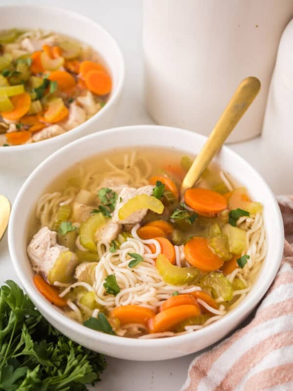 A bowl of slow cooker chicken soup with sliced carrots, celery, tender chicken pieces, and parsley is served with a gold spoon next to fresh parsley and a striped cloth napkin. Another bowl of comforting soup appears in the background.