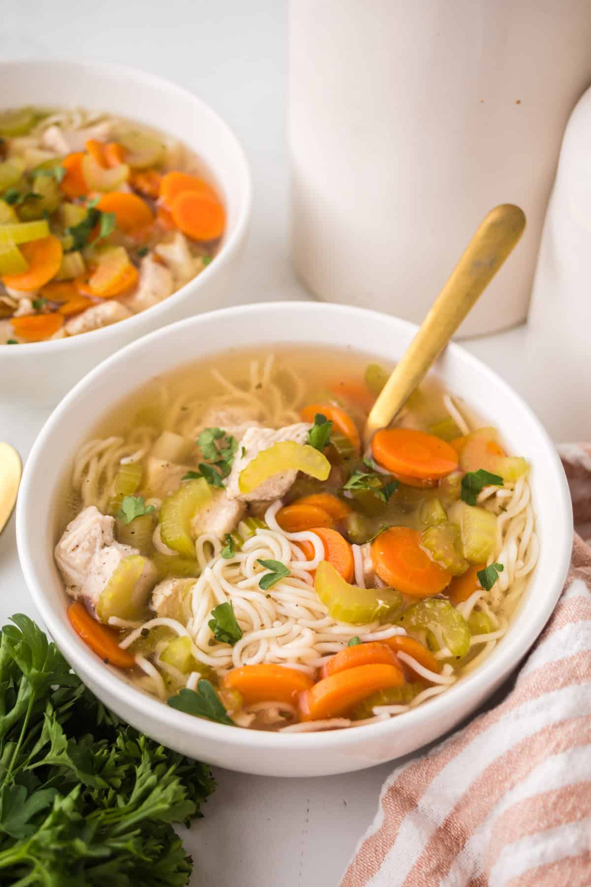 A bowl of slow cooker chicken soup with sliced carrots, celery, tender chicken pieces, and parsley is served with a gold spoon next to fresh parsley and a striped cloth napkin. Another bowl of comforting soup appears in the background.